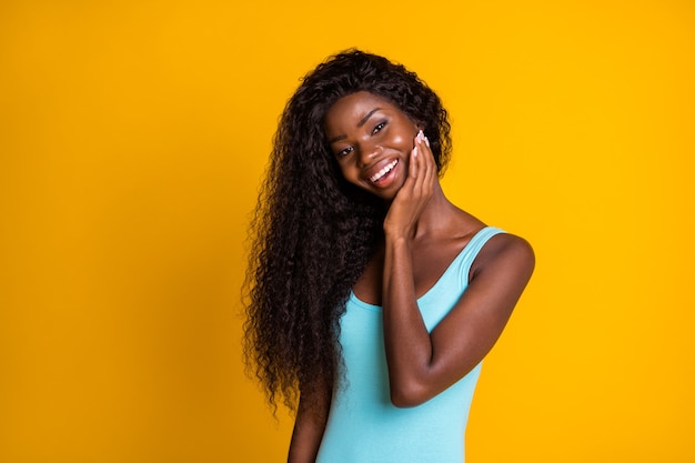 Portrait photo d'une charmante fille afro-américaine avec une peau parfaite touchant le visage avec un sourire à la main isolé sur un fond de couleur jaune vif