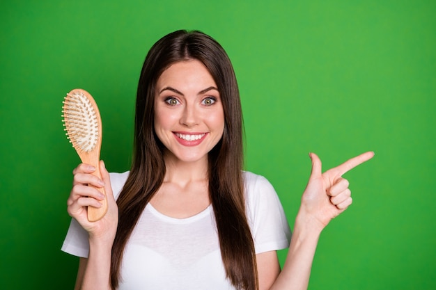 Portrait photo d'une femme pointant le doigt sur le côté tenant une brosse à cheveux à la main isolée sur fond de couleur vert vif