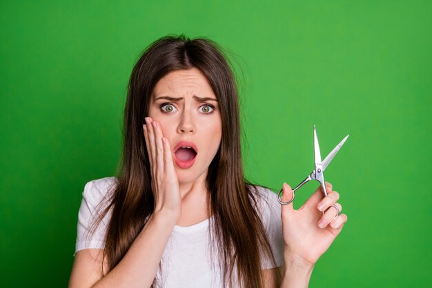 Portrait Photo D'une Fille Bouleversée Tenant Des Ciseaux Touchant Le Visage Avec Une Bouche Ouverte D'une Main Isolée Sur Fond De Couleur Vert Vif