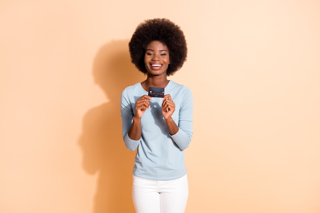 Portrait photo d'une fille joyeuse et heureuse à la peau foncée montrant une carte bancaire en plastique portant une chemise bleue isolée sur fond de couleur beige