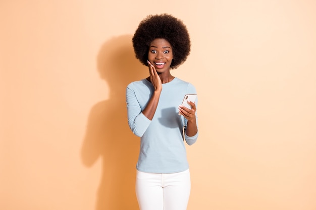 Portrait photo d'une fille à la peau foncée surprise heureuse tenant un smartphone touchant le visage avec la main isolée sur fond de couleur beige