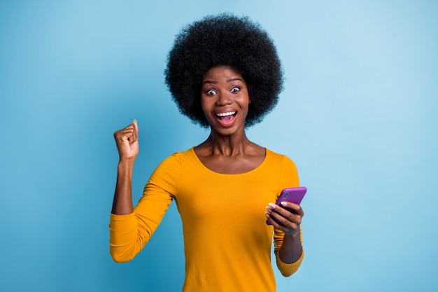Portrait photo d'une fille à la peau noire étonnée faisant des gestes comme un gagnant en gardant le smartphone isolé sur un fond de couleur bleu vif