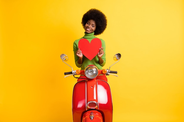 Portrait photo d'une fille à la peau noire sur une moto tenant une carte postale grand coeur rouge à deux mains isolée sur fond de couleur jaune vif