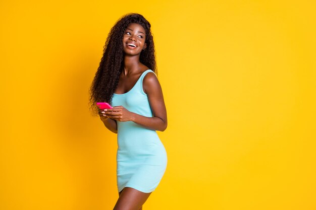 Portrait Photo D'une Influenceuse Brune Afro-américaine Tenant Un Téléphone Rose Avec Deux Mains Riant Vêtue D'une Mini-robe Bleue Isolée Sur Fond De Couleur Jaune Vif Avec Fond