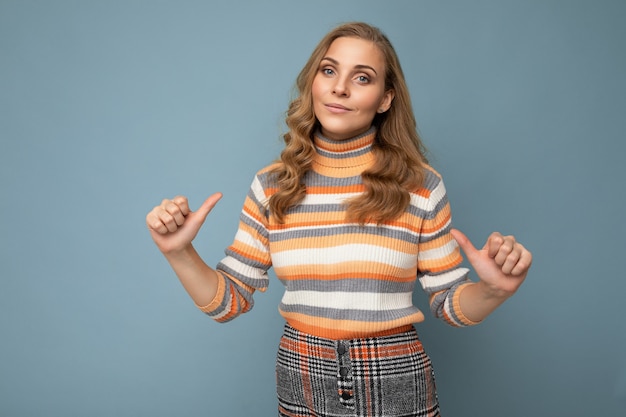 Portrait photo d'une jeune femme blonde assez belle et confiante avec des émotions sincères portant un pull rayé isolé sur fond bleu avec un espace vide et se pointant du doigt.