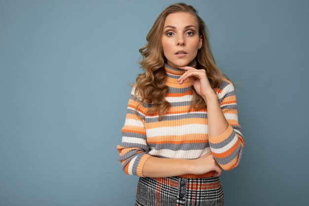 Portrait photo d'une jeune femme blonde réfléchie assez belle portant un élégant pull rayé coloré isolé sur fond bleu avec espace de copie et réflexion.