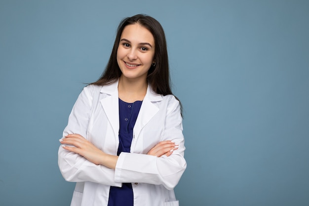 Portrait de photo de jeune femme brune souriante positive assez belle avec des émotions sincères portant une blouse médicale blanche isolée sur fond bleu avec espace copie et tenant les bras croisés.