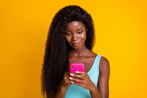 Portrait photo d'une jeune fille afro-américaine tenant un téléphone rose à deux mains portant un débardeur bleu isolé sur un fond de couleur jaune vif