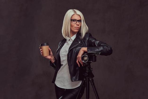 Portrait d'une photographe blonde intelligente dans des vêtements et des lunettes à la mode tient un café à emporter et pose tout en s'appuyant sur un appareil photo professionnel avec un trépied dans un studio, regarde l'appareil photo. je