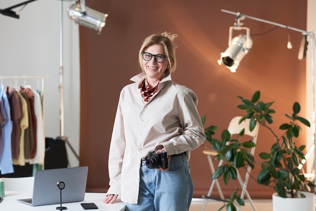 Portrait de photographe de mode mature souriant à la caméra en se tenant debout au studio avec appareil photo