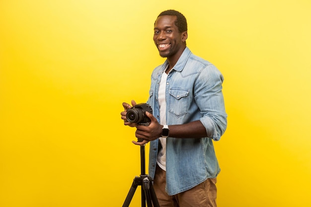 Portrait d'un photographe motivé excité ou d'un voyageur en chemise décontractée en denim souriant et posant à la caméra avec un appareil photo reflex numérique professionnel sur un trépied tourné en studio isolé sur fond jaune