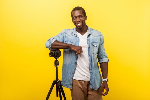 Portrait d'un photographe ou d'un voyageur joyeux en chemise décontractée en jean souriant et posant à l'appareil photo avec un appareil photo reflex numérique professionnel sur trépied. studio d'intérieur tourné isolé sur fond jaune