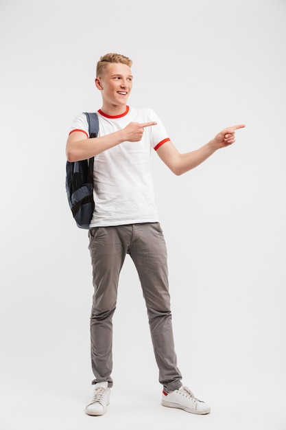 Portrait en pied d'étudiant garçon portant des vêtements décontractés et sac à dos, pointant le doigt de côté au fond, isolé sur blanc