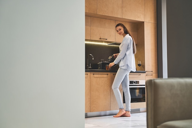 Portrait en pied d'une femme souriante versant de l'eau d'une bouilloire électrique dans une tasse