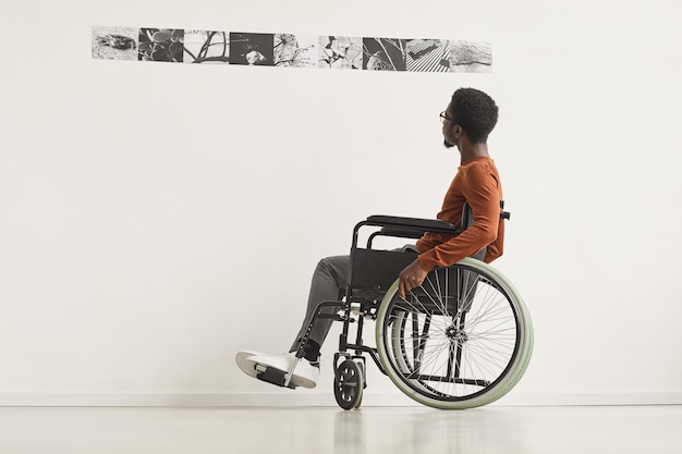 Portrait en pied graphique de jeune homme afro-américain utilisant un fauteuil roulant et regardant des peintures tout en explorant l'exposition de la galerie d'art moderne,