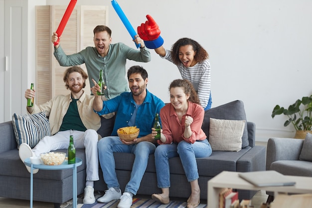 Portrait en pied d'un groupe multiethnique d'amis regardant un match de sport à la télévision et applaudissant émotionnellement assis ensemble sur un canapé