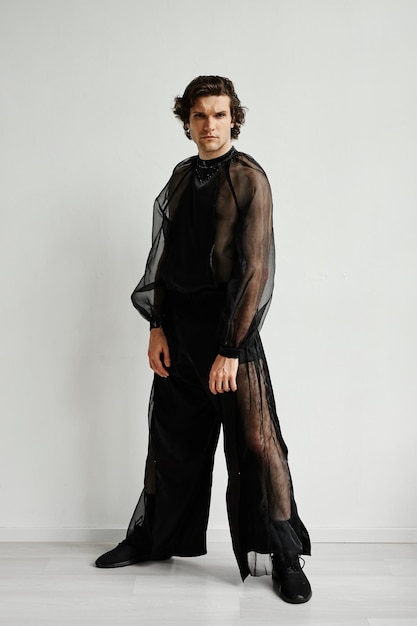 Portrait en pied d'un homme non binaire portant une tenue en mousseline de soie noire posant contre le blanc à la mode