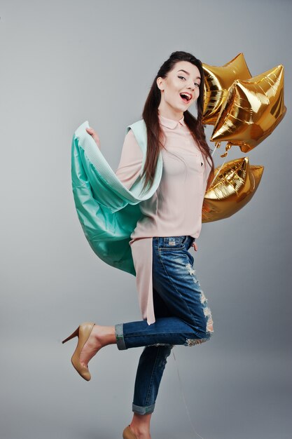 Portrait en pied jeune fille brune vêtue d'une blouse rose, d'une veste turquoise, de jeans déchirés et de chaussures crème tenant des ballons d'étoiles dorées.