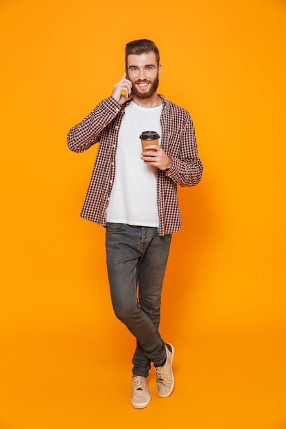 Portrait en pied d'un jeune homme gai portant des vêtements décontractés tenant une tasse de café à emporter parler au téléphone mobile