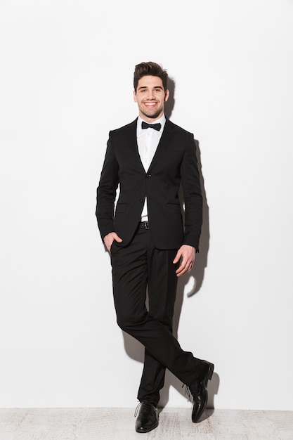 Portrait en pied d'un jeune homme souriant vêtu d'un costume noir avec un arc debout isolé sur blanc, posant