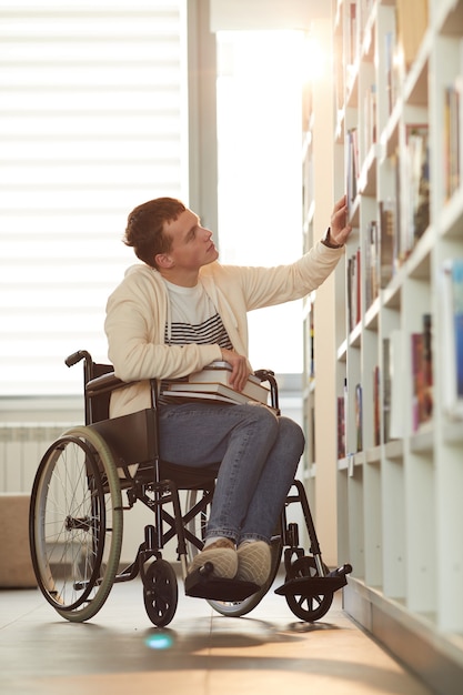 Portrait en pied vertical de jeune homme utilisant un fauteuil roulant à l'école tout en regardant des étagères dans la bibliothèque éclairée par la lumière du soleil