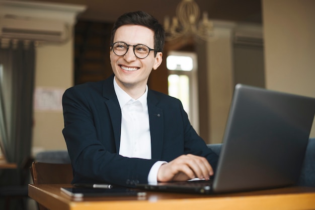 Portrait D'un Pigiste Adulte Attrayant Portant Costume Et Lunettes à La Voiture En Souriant Tout En Travaillant Sur Son Ordinateur Portable Assis Dans Un Café.