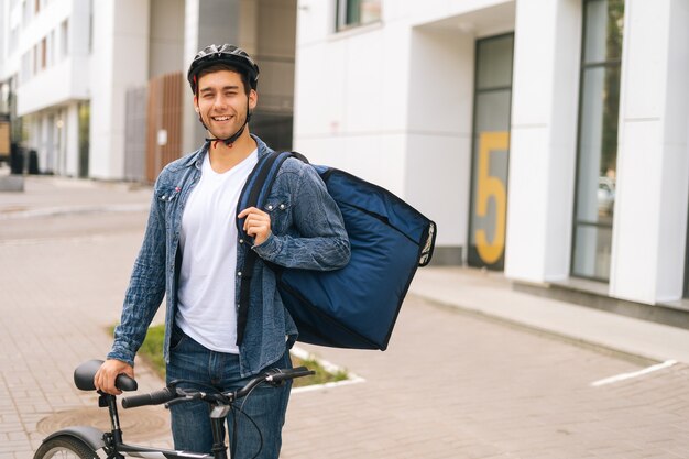 Portrait de plan moyen d'un beau jeune livreur joyeux dans un casque de protection posant debout près d'un vélo dans la rue de la ville, regardant la caméra. Courier mâle avec sac à dos livraison de nourriture au client