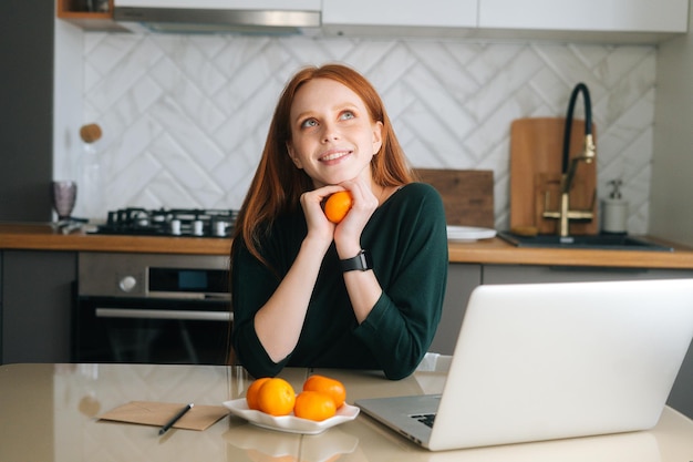 Portrait de plan moyen d'une jeune femme rousse joyeuse assise à table avec un ordinateur portable et tenant