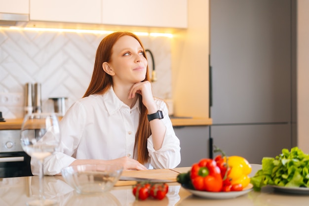 Portrait de plan moyen d'une jeune femme rousse rêvante assise à la table de la cuisine avec une planche à découper