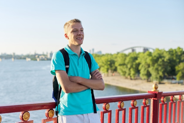 Portrait en plein air d'un adolescent souriant de 15, 16 ans, les bras croisés. Mâle sur le pont au-dessus de la rivière aux beaux jours d'été