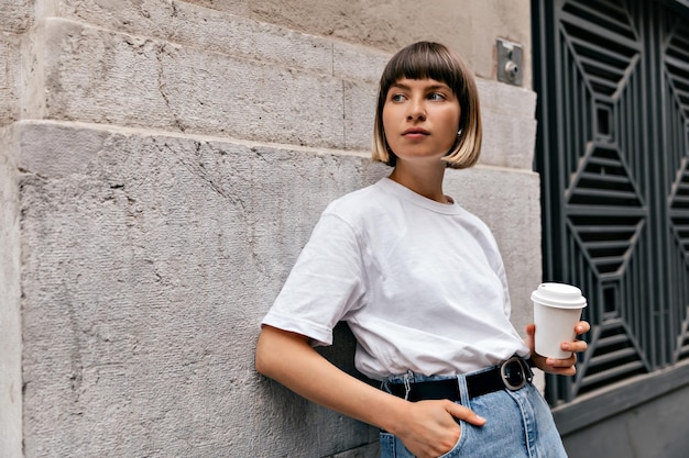 Portrait en plein air d'une adolescente élégante avec une coiffure courte portant un t-shirt blanc et un jean tient un café et regarde de côté tout en marchant dans la ville par temps chaud