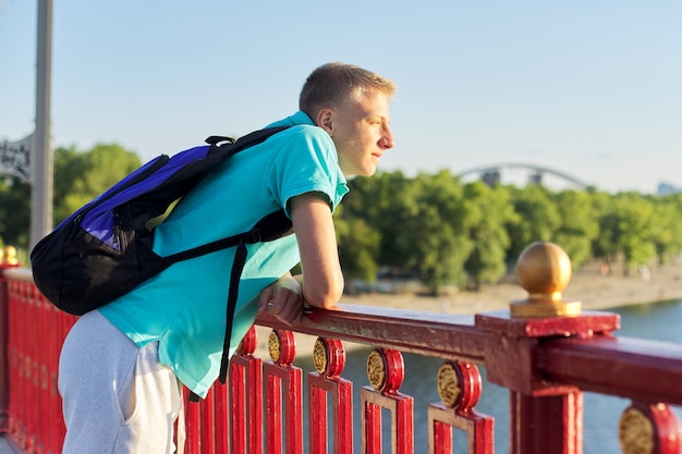 Portrait en plein air d'un beau garçon adolescent de 15, 16 ans, avec espace de copie. Guy blond avec sac à dos regarde la rivière, debout sur le pont, coucher de soleil journée d'été ensoleillée, mode de vie urbain