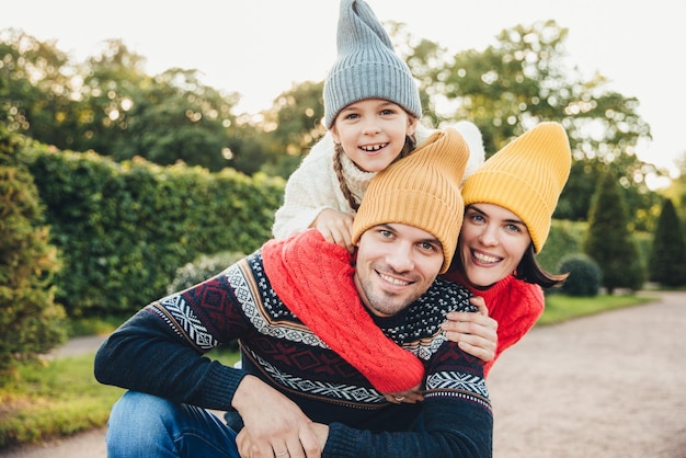 Portrait en plein air d'un bel homme donner le dos à sa femme et sa fille porter des vêtements chauds avoir des expressions heureuses se soutenir mutuellement Famille embrasser à l'extérieur sourire agréablement devant la caméra