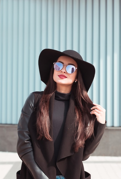 Portrait en plein air d'une belle mannequin brune portant un manteau noir et un chapeau à larges bords. Concept de mode féminine