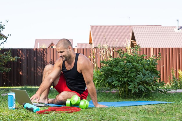 Le portrait en plein air du jeune homme en uniforme de sport se repose, s'étend sur la pelouse, regarde un film et étudie depuis un ordinateur portable, un réseau social dans le jardin