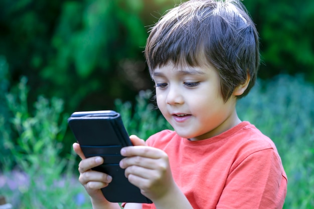 Portrait en plein air enfant avec un visage heureux jouant à un jeu sur téléphone mobile, mignon garçon s'amusant à regarder des dessins animés sur téléphone, enfant avec visage souriant jouant à des jeux.