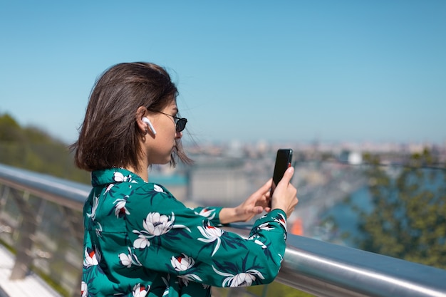 Portrait en plein air d'une femme en chemise verte décontractée à une journée ensoleillée se dresse sur le pont à la recherche sur l'écran du téléphone des écouteurs bluetooth sans fil dans les oreilles