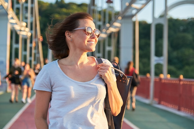 Portrait en plein air d'une femme mature souriante heureuse dans des lunettes de soleil marchant sur le pont, heure d'or, saison estivale