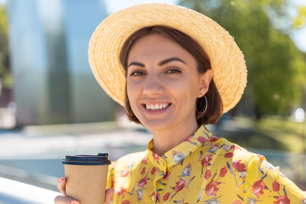 Portrait en plein air de femme en robe d'été jaune et chapeau avec tasse de café en profitant du soleil