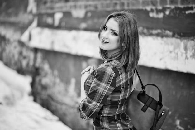 Un portrait en plein air d'une jeune jolie fille aux cheveux rouges portant une robe à carreaux avec des sacs à dos girly debout sur le fond du mur de briques en hiver Photo en noir et blanc