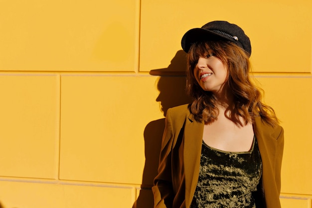 Portrait en plein air de jolie femme souriante avec une coiffure frisée portant veste et casquette posant sur mur jaune