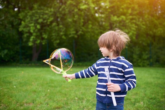 Portrait en plein air de mignon garçon d'âge préscolaire soufflant des bulles de savon sur une pelouse verte à l'aire de jeux.