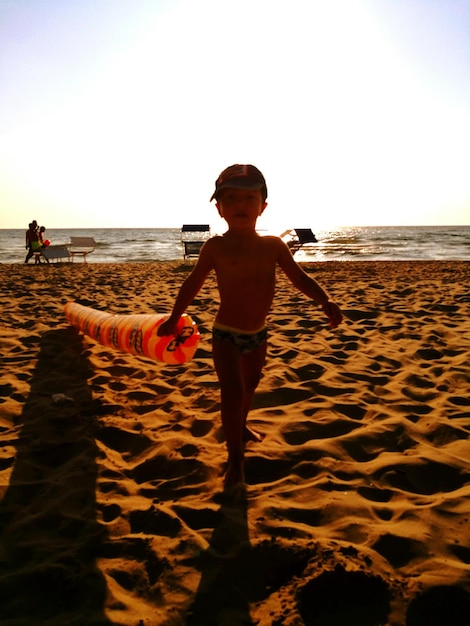 Photo portrait en pleine longueur d'un bébé garçon avec un jouet debout sur le sable à la plage contre un ciel clair