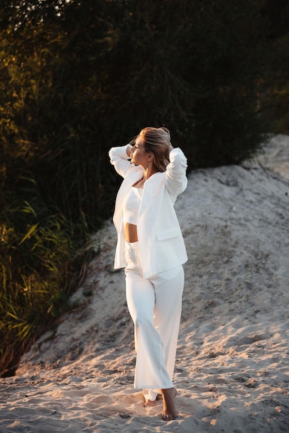 Portrait en pleine longueur d'une belle jeune femme aux cheveux blonds debout pieds nus sur le sable tout en profitant d'un magnifique coucher de soleil Modèle de mode portant un costume blanc à la mode