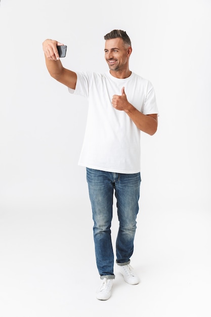 Portrait de pleine longueur d'un homme souriant des années 30 portant un t-shirt décontracté et un jean prenant une photo de selfie sur un téléphone portable tout en tenant dans la main isolé sur blanc