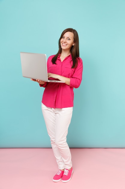 Portrait de pleine longueur jeune femme en chemise rose chemisier, pantalon blanc tenant un ordinateur portable isolé sur un mur pastel bleu rose vif.