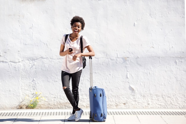 Portrait pleine longueur de jeune femme noire debout avec des bagages à l&#39;extérieur et souriant