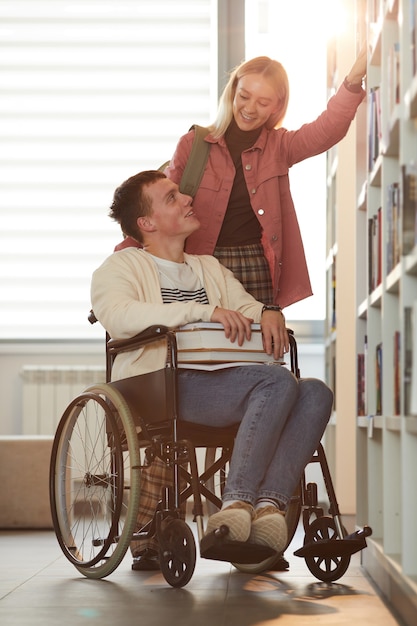 Portrait De Pleine Longueur Verticale De Jeune Homme Utilisant Un Fauteuil Roulant à L'école Avec Une Amie L'aidant Dans La Bibliothèque éclairée Par La Lumière Du Soleil