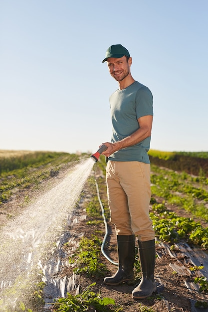 Portrait de pleine longueur verticale de jeune travailleur masculin arrosant les cultures à la plantation de légumes et souriant à la caméra tout en se tenant à l'extérieur contre le ciel bleu