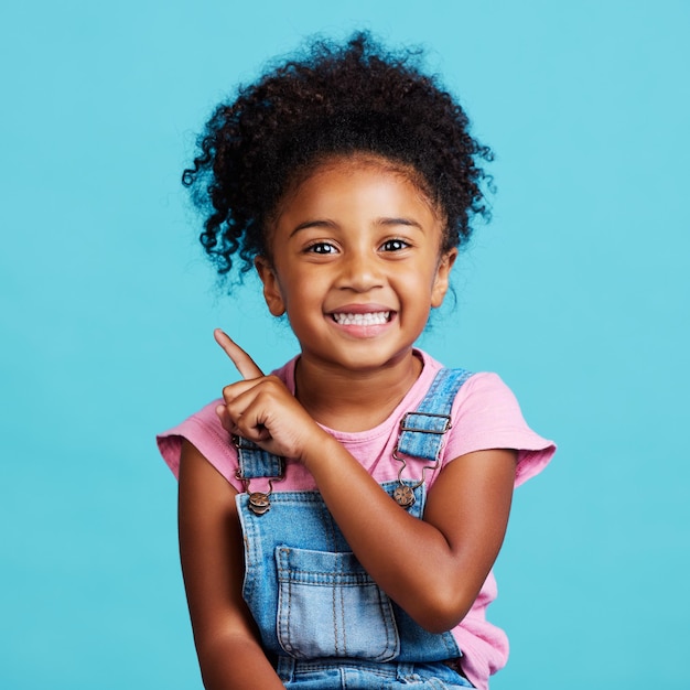 Portrait pointant et maquette avec une fille sur fond bleu en studio montrant l'espace de placement de produit Marketing et publicité pour enfants avec une adorable enfant de sexe féminin à l'intérieur pour pointer vers l'image de marque